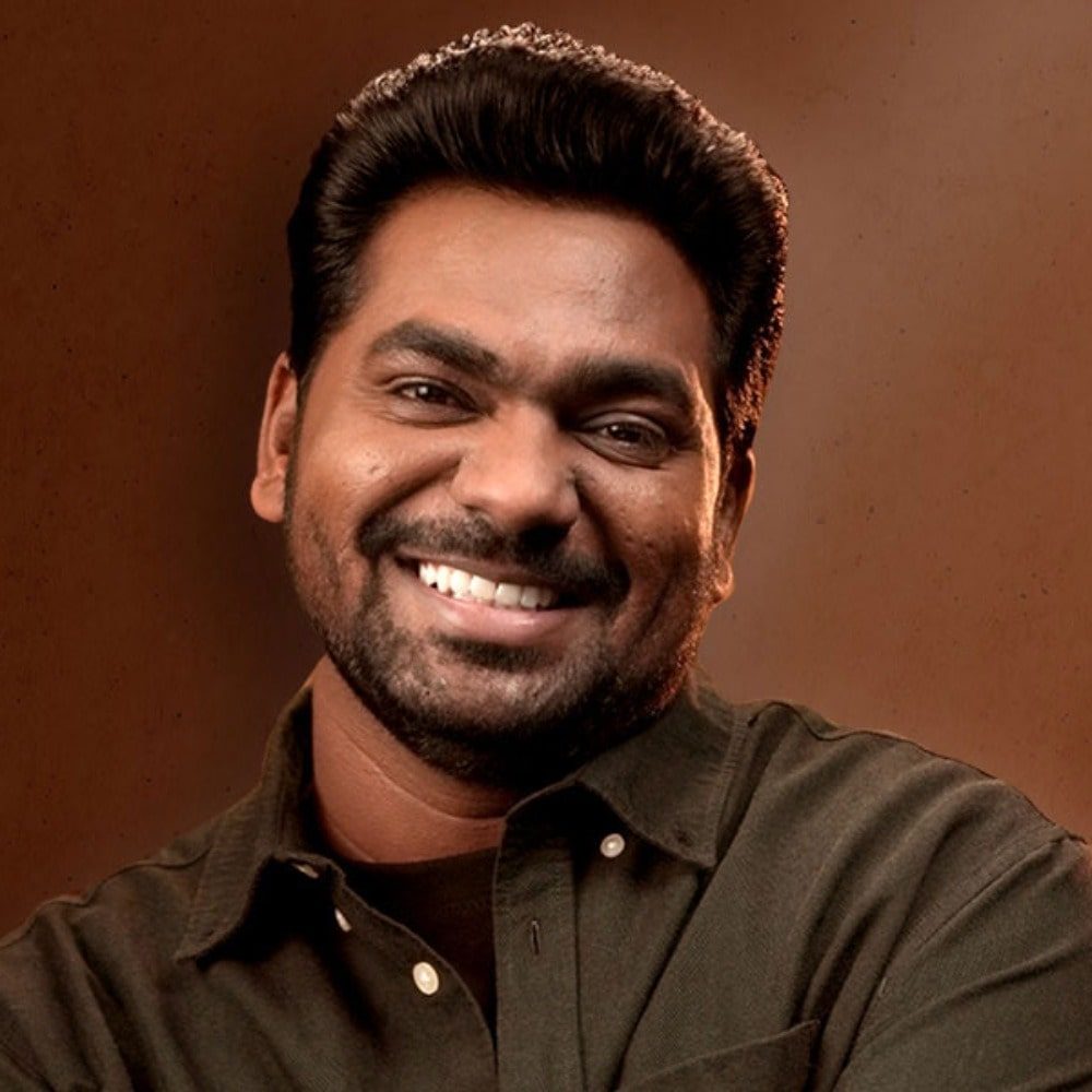 A smiling man with a neatly groomed beard and hair, resembling Zakir Khan, wearing a dark shirt against a brown background.