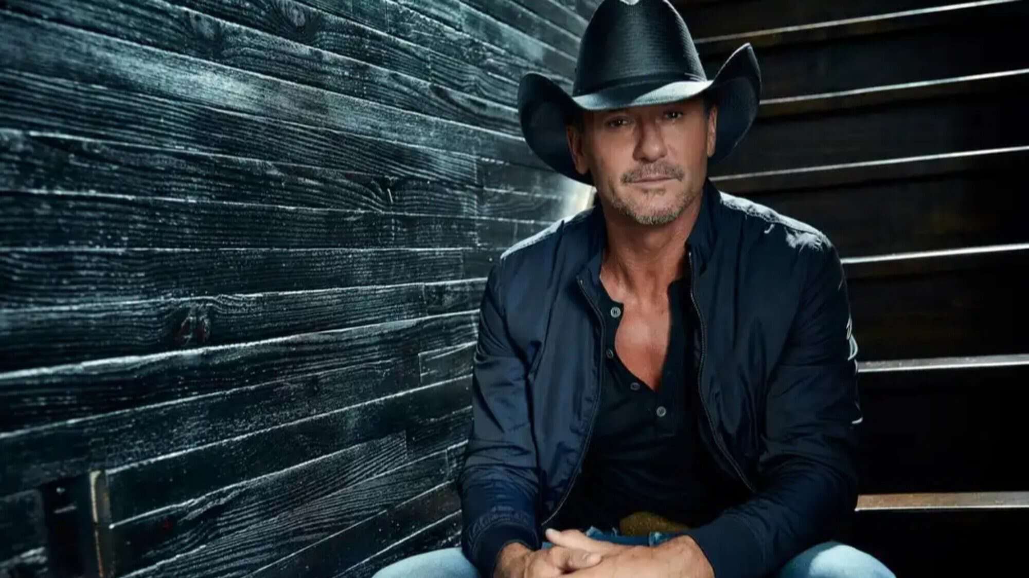 Tim McGraw in cowboy hat sitting on steps against a black wood background.