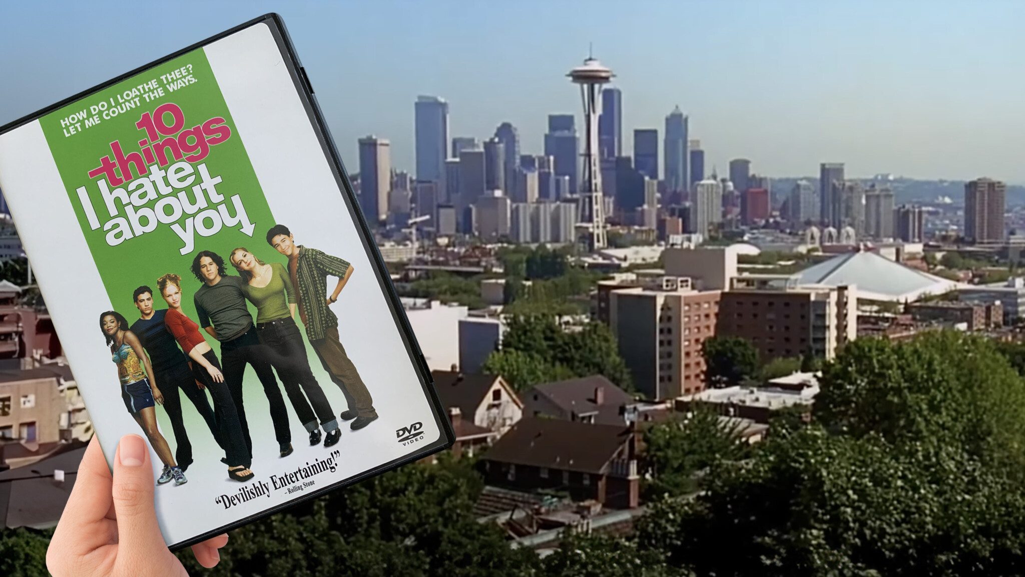 Hand holding a DVD of "10 Things I Hate About You" in front of a city skyline that matches the setting of the movie, celebrating its 25th anniversary.