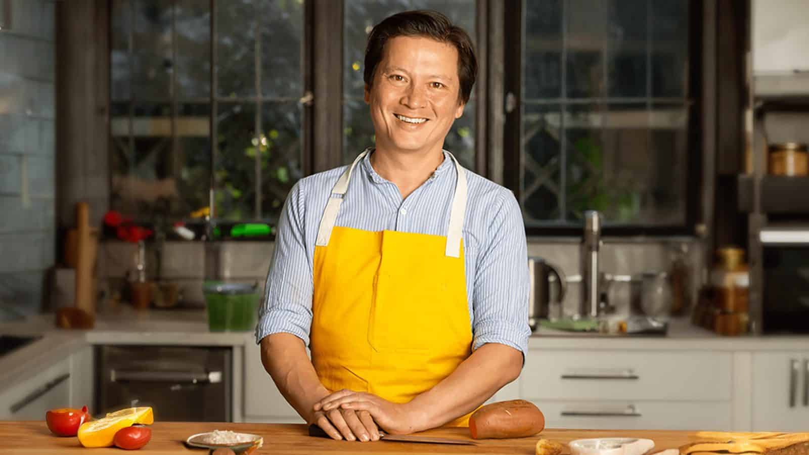 A smiling person wearing a blue-striped shirt and a yellow apron stands at a kitchen counter, engaged in Kitchen Conversations with various food items, inspired by J. Kenji López-Alt.