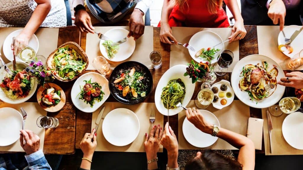 Overhead view of a dinner table with six people dining on various meals curated by Chef Luke Kolpin, including salads, seafood, and vegetables, with wine and water glasses.