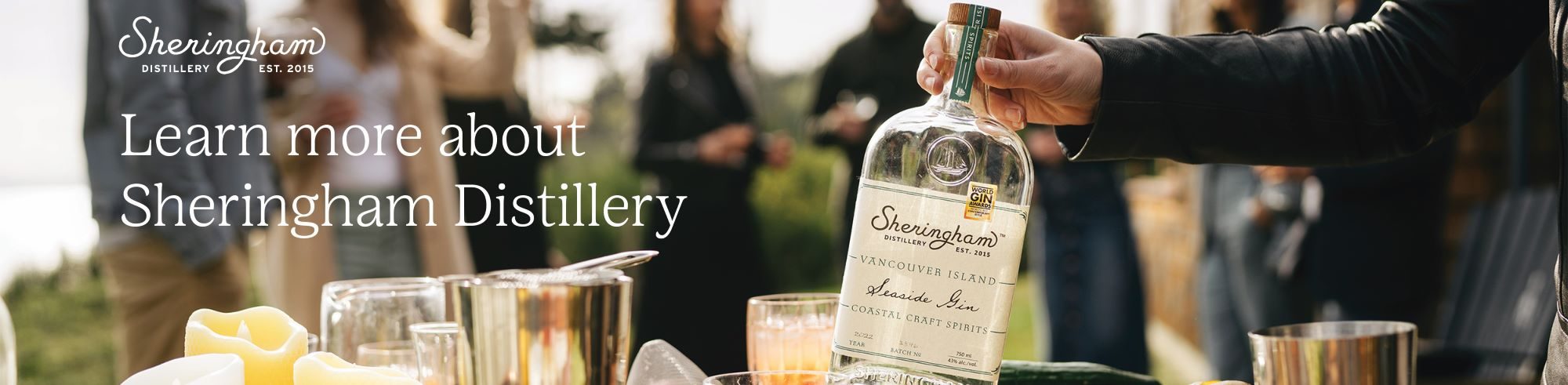 A person pours Sheringham Distillery gin into a glass at a culinary retreat with people chatting in the background.