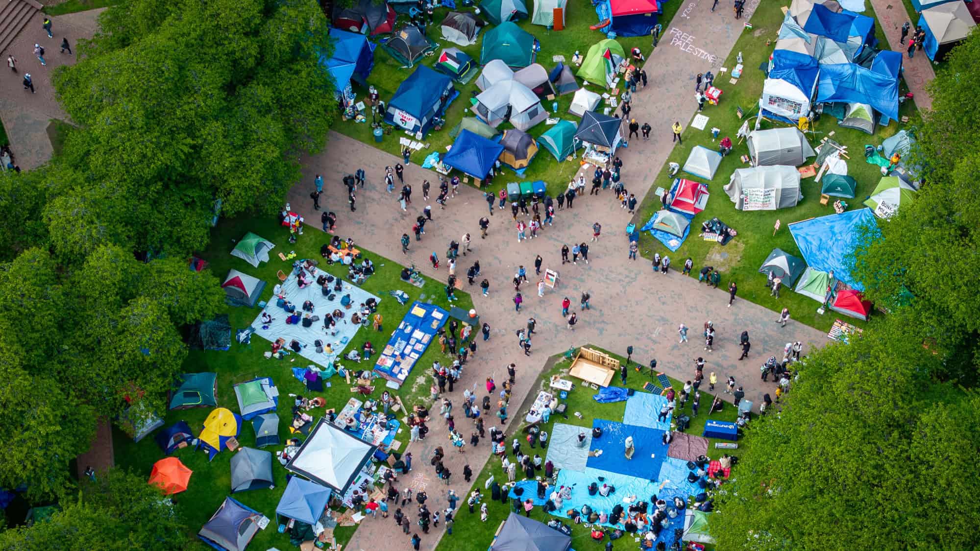 An aerial view of a park with people gathering around various tents and blue tarps in different sections, bordered by green trees. The scene suggests an outdoor event or festival, reminiscent of an encampment deal fostering a range of opinions and ideas.