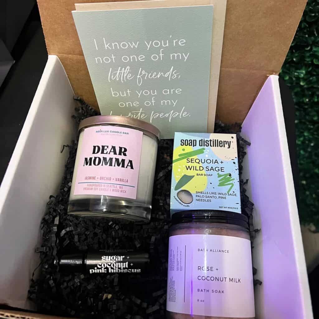 Gift box containing various Mother's Day self-care products including scented candles, soap, bath soak, and lip balm, alongside a card with a heartfelt message.