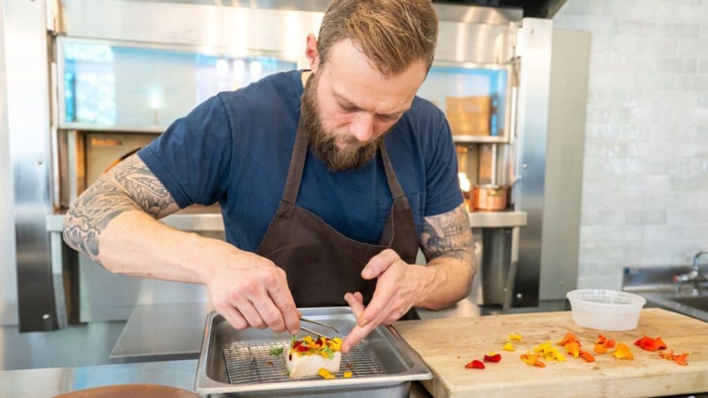 Chef Luke Kolpin, a bearded chef with tattoos, garnishes a dish with edible flowers in a professional kitchen.