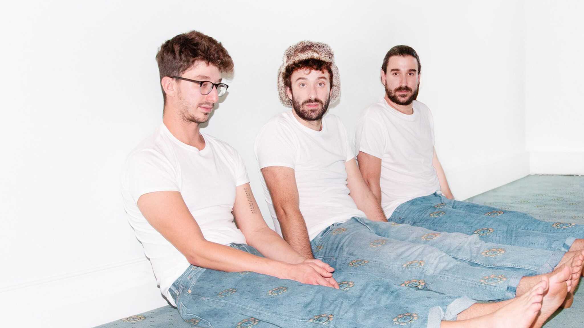 Three men in white t-shirts and jeans sitting side by side against a white wall, looking at the camera, possibly from the AJR Maybe Man Tour.