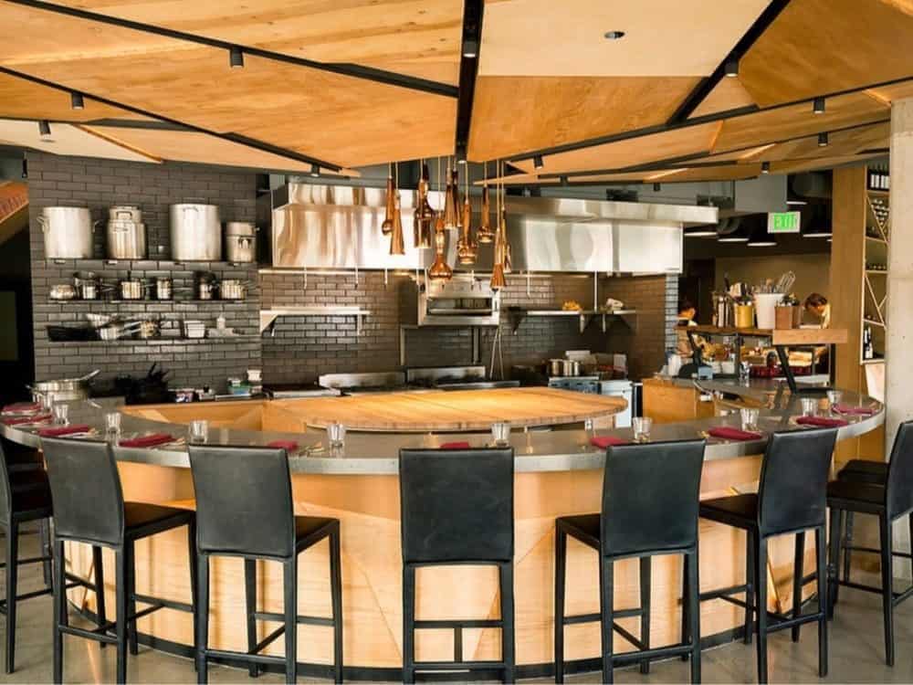 Modern culinary retreat interior showing an open kitchen, bar stools in front, hanging copper pots, and a wooden ceiling.