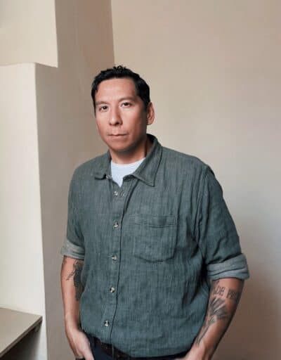 Portrait of artist Sky Hopinka wearing a green shirt and several tattoos on his arms