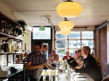 A bartender serves customers at a bar with large, round hanging lights and a brick wall in the background, featuring spirits from Sheringham Distillery.