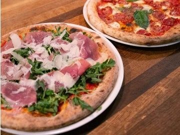 Two pizzas on wooden tables; one topped with prosciutto, arugula, and shaved parmesan, the other with tomato, mozzarella, and basil at a culinary retreat.