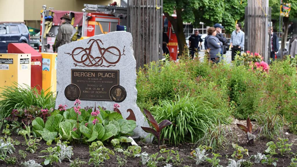 Image of Bergen Place, a memorial stone in a garden setting with people and emergency vehicles in the background. The stone features inscriptions and circular plaques, capturing the lively atmosphere as locals celebrate Norwegian Constitution Day with chants of "hip hip hip hurra.