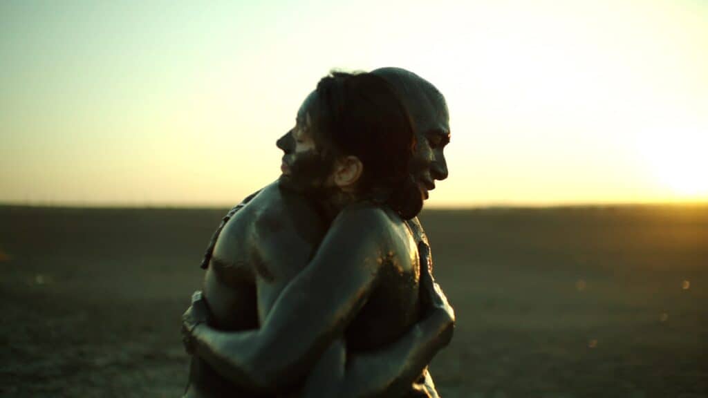 Two individuals covered in mud embrace each other in a desert landscape at sunset.