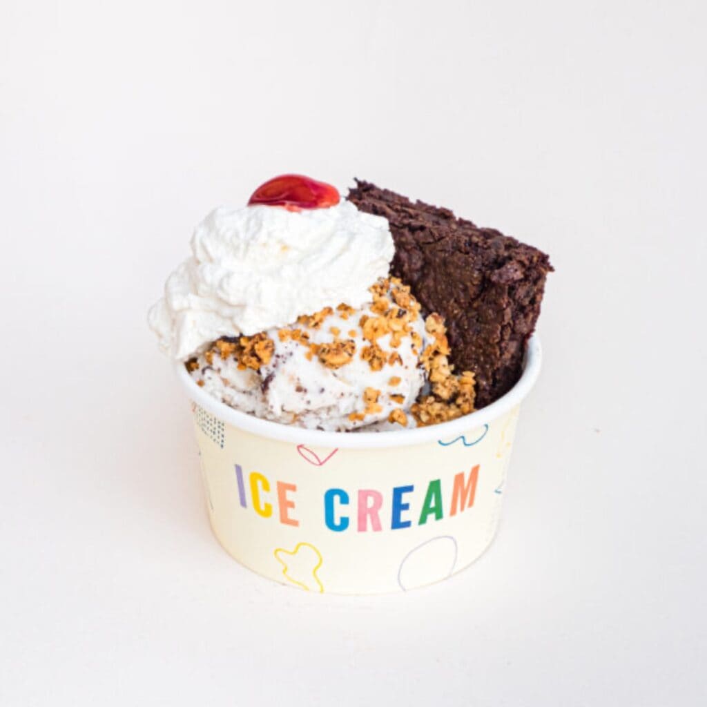 A bowl of ice cream topped with whipped cream, a cherry, popcorn, and a brownie, perfect for Mother's Day, against a plain white background.