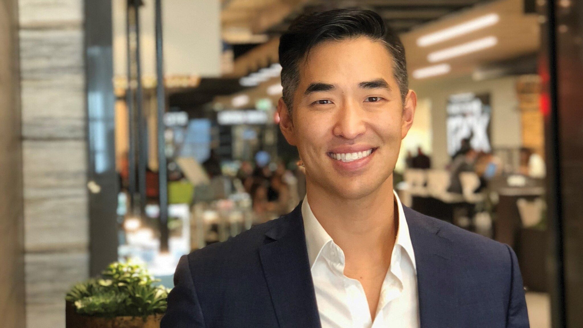 A smiling man in business attire posing in a busy indoor setting, representing Uncommon Thinkers.