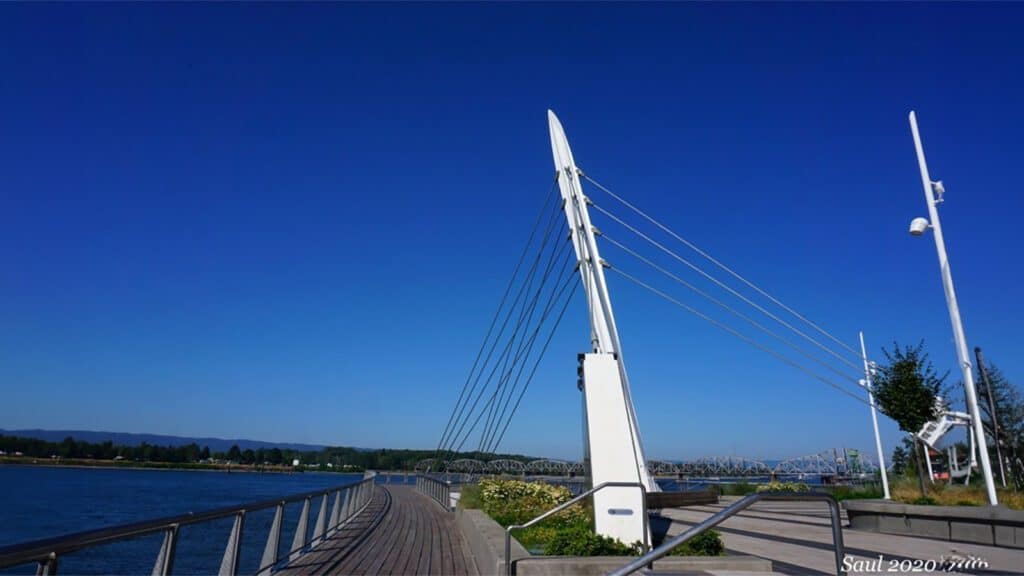 A modern cable-stayed bridge with a white pylon over a wooden boardwalk by a river in Vancouver Washington, under a clear blue sky.