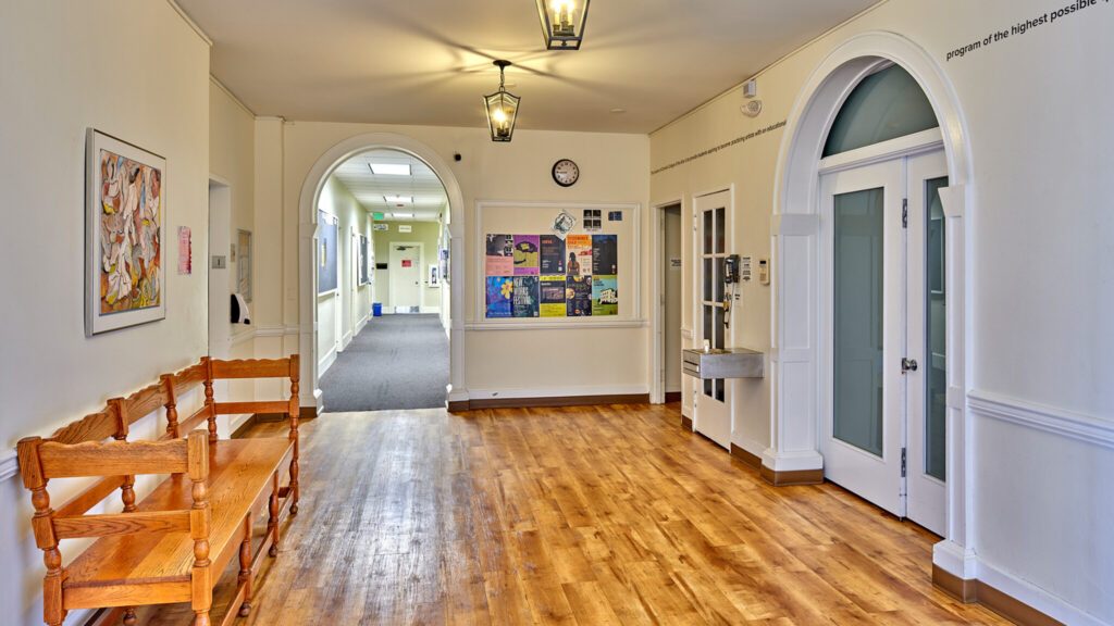 Brightly lit hallway in a building with wooden benches, artwork on the walls, and a water fountain.