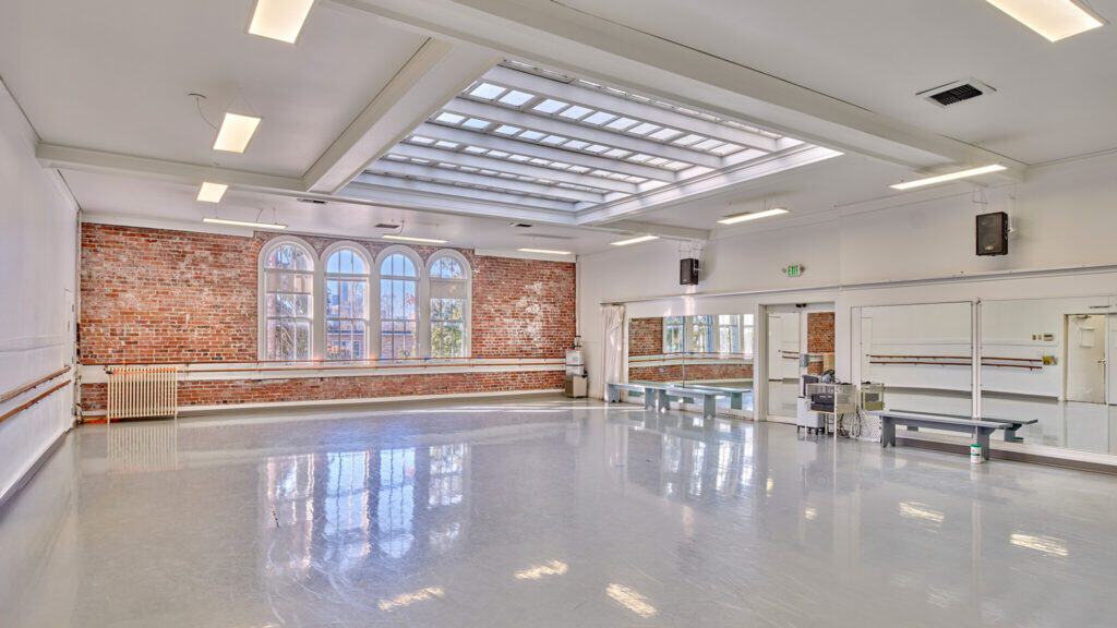 An empty dance studio with large mirrors, a glossy floor, and exposed brick walls featuring large arched windows.