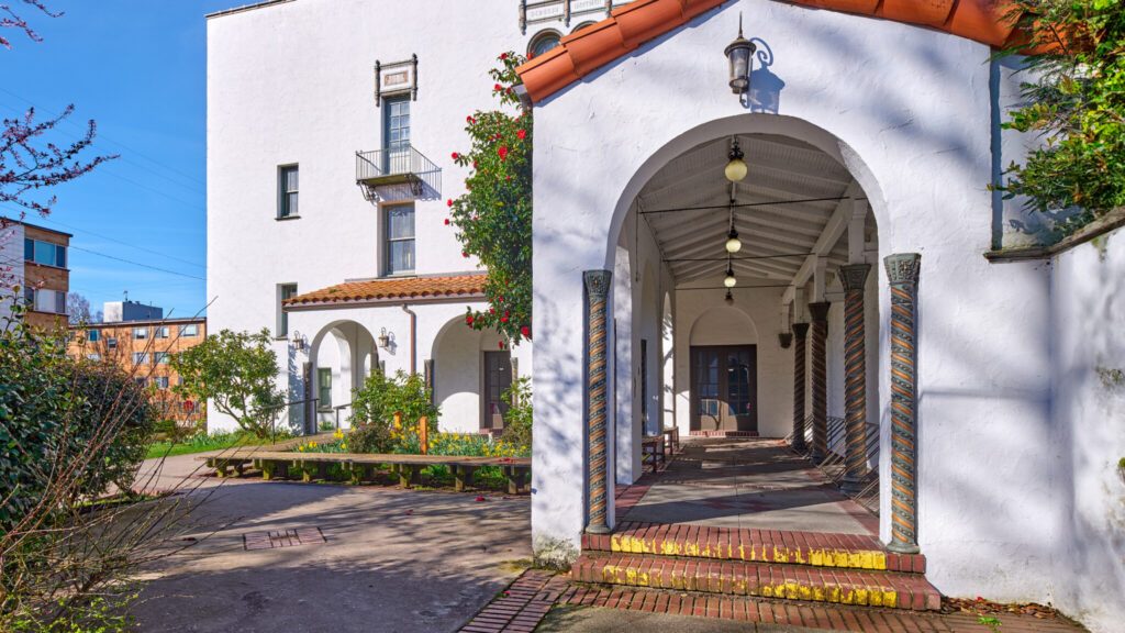 A sunny courtyard with a covered walkway featuring decorative columns, adjacent to a multi-story white building.