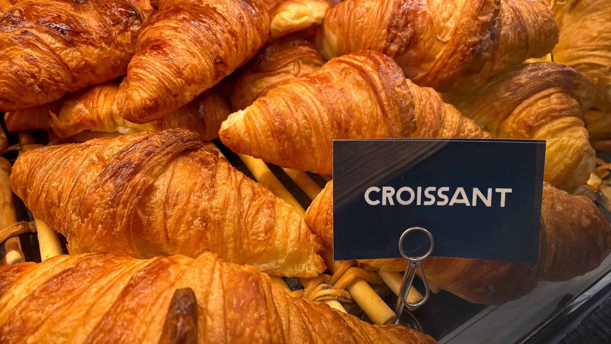 A display of golden-brown croissants with a black sign labeled "croissant" (pronunciation: kruh-sahnt) in a bakery case.