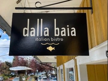 Sign for "dalla baia italian bistro" with a gold bird icon, hanging outside a building with yellow trim, featured in Seattle magazine.