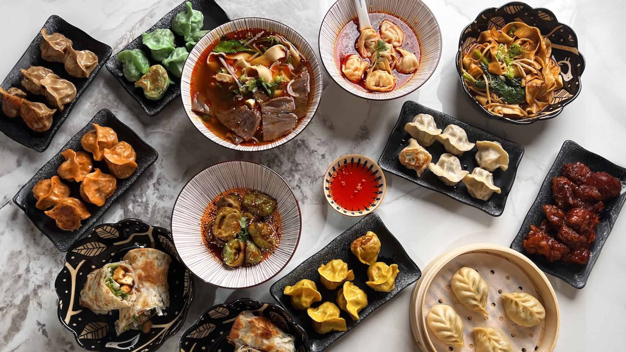 Assorted Asian dumplings and noodle dishes displayed on various patterned plates, with dipping sauces, featured during Seattle Restaurant Week, on a marble surface.