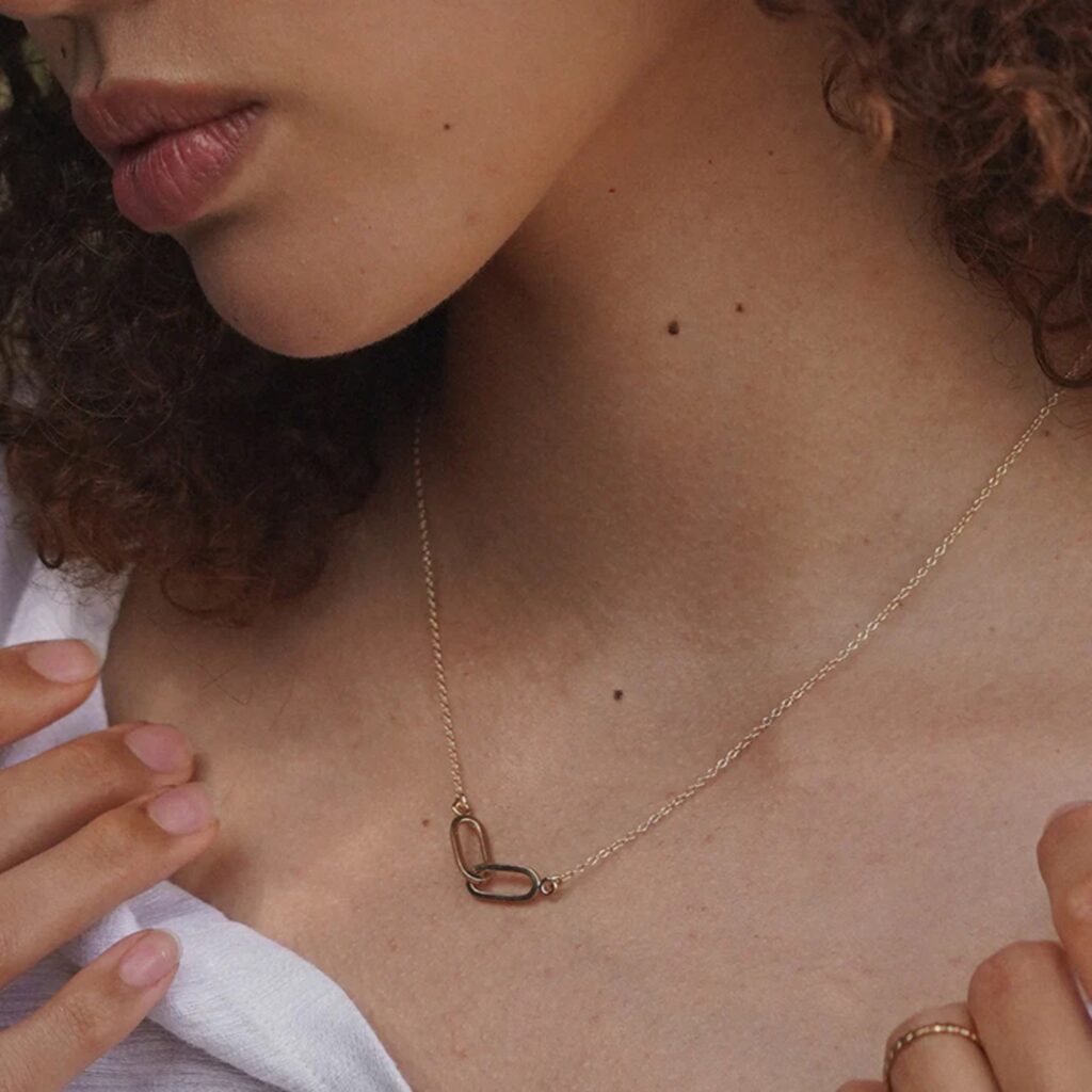 Close-up of a woman with curly hair wearing a delicate gold necklace, the perfect Mother's Day gift, featuring a pendant resembling an infinity symbol.