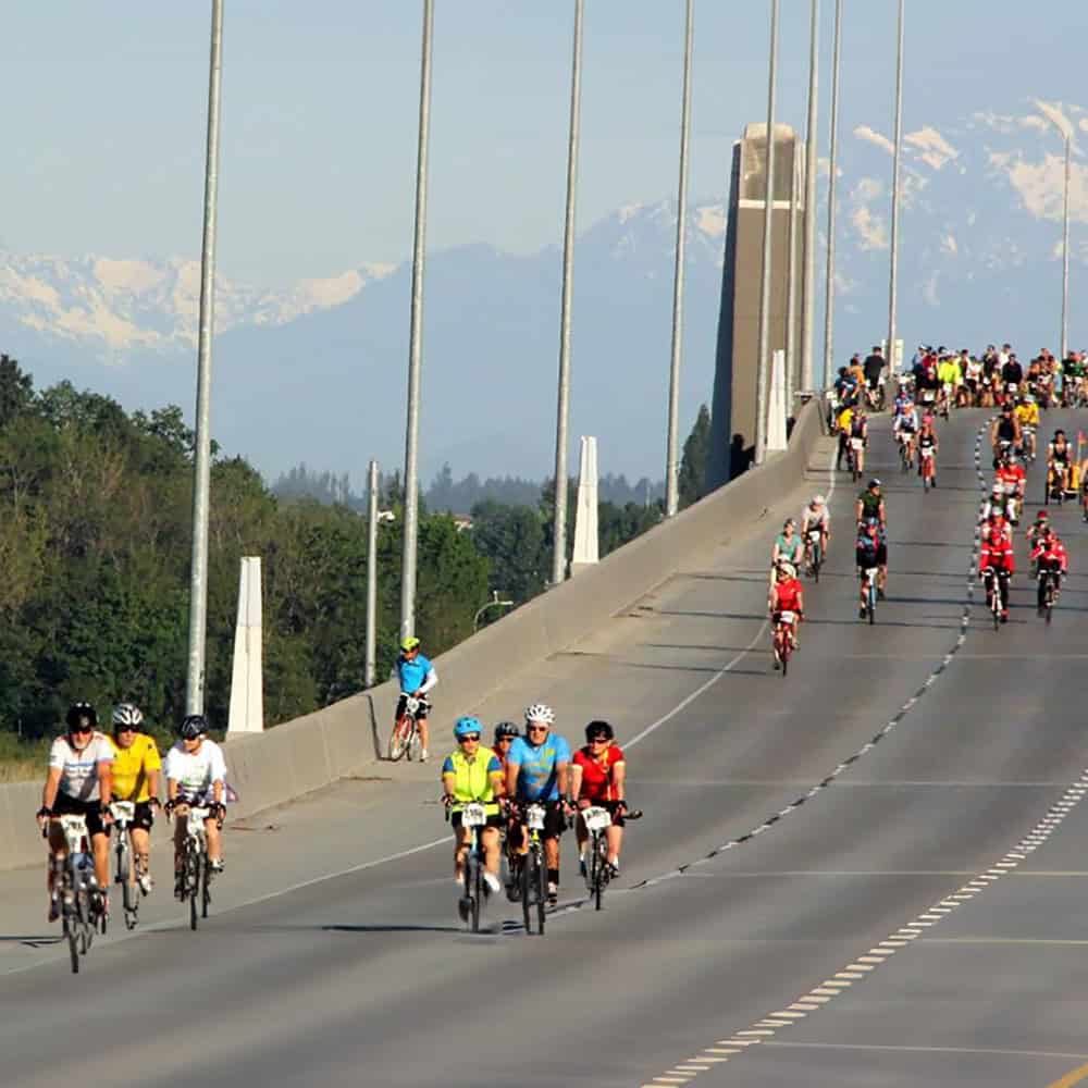 Group of cyclists riding on a bridge with mountains in the background on a sunny day during the "Must List" week of May 2-8.