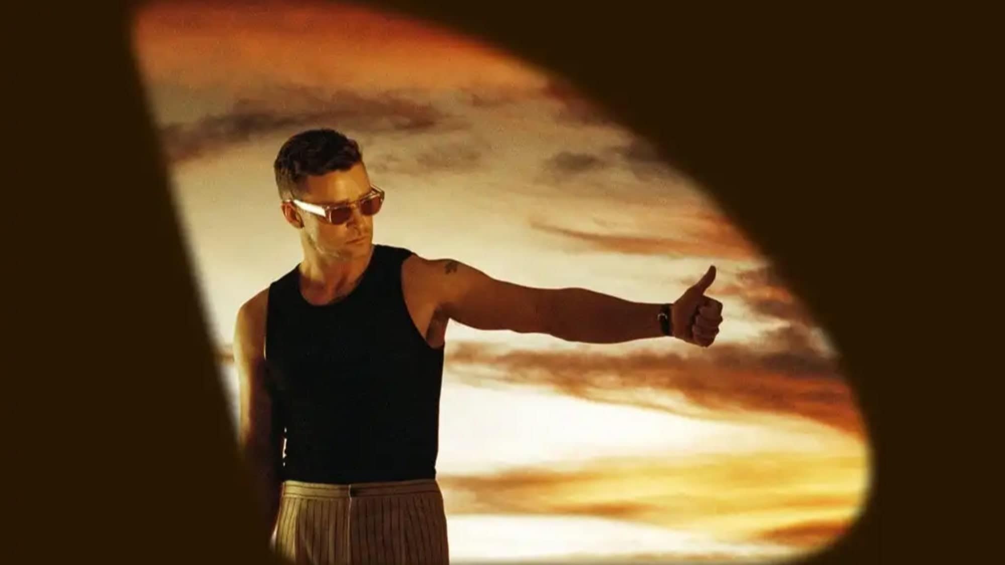 Man in sunglasses giving a thumbs up, viewed through a sunglasses lens against a sunset background at the Justin Timberlake concert tour.