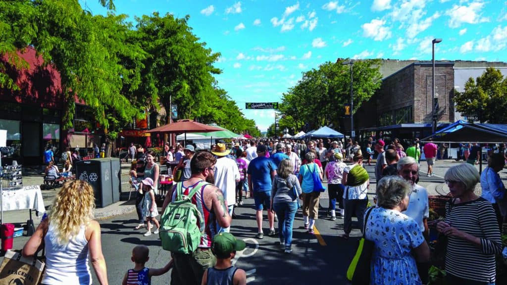 Crowded street festival in Iconic Idaho with people browsing vendor stalls under sunny skies, flanked by trees and buildings.