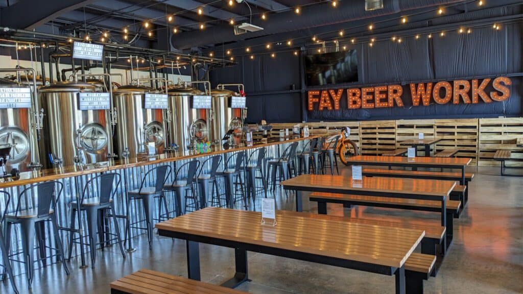 Interior of a brewery with stainless steel tanks, a long bar with stools, and wooden benches under a sign reading "Pioneer Square Fay Beer Works.