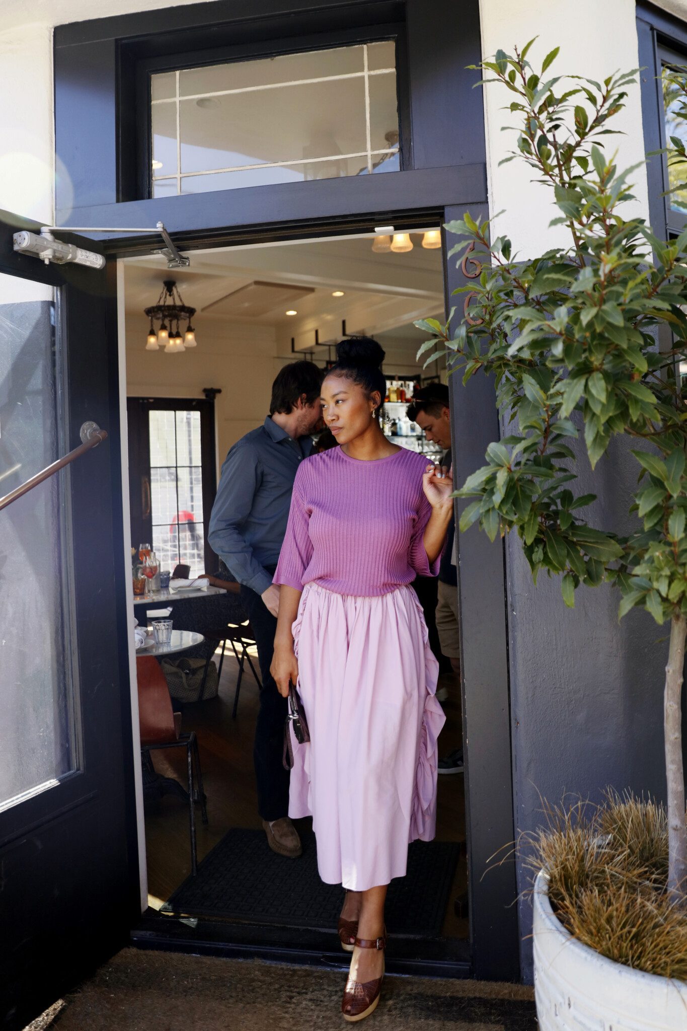 A person in a dress stands indoors near a window, holding a houseplant in a flowerpot, while a person stands outside the door of a building, admiring the plant.