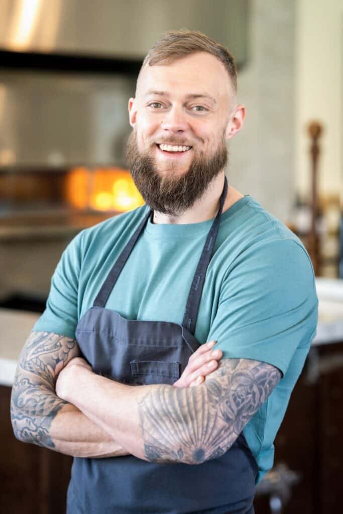 portrait of a Chef Luke Kolpin standing in front of the Cedar and Elm kitchen with his arms crossed and wearing a dark blue apron.