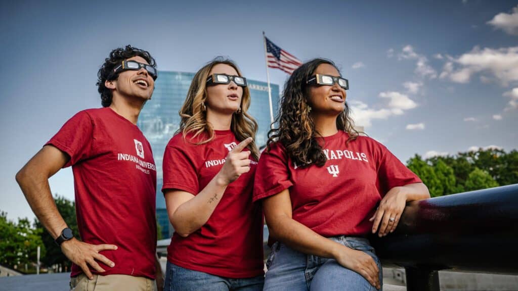 Three individuals in matching red t-shirts and sunglasses observing a solar eclipse, looking away from the camera with a cityscape in the background.