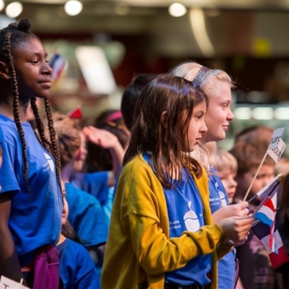 Group of children attentively participating in a Must List event, holding flags.