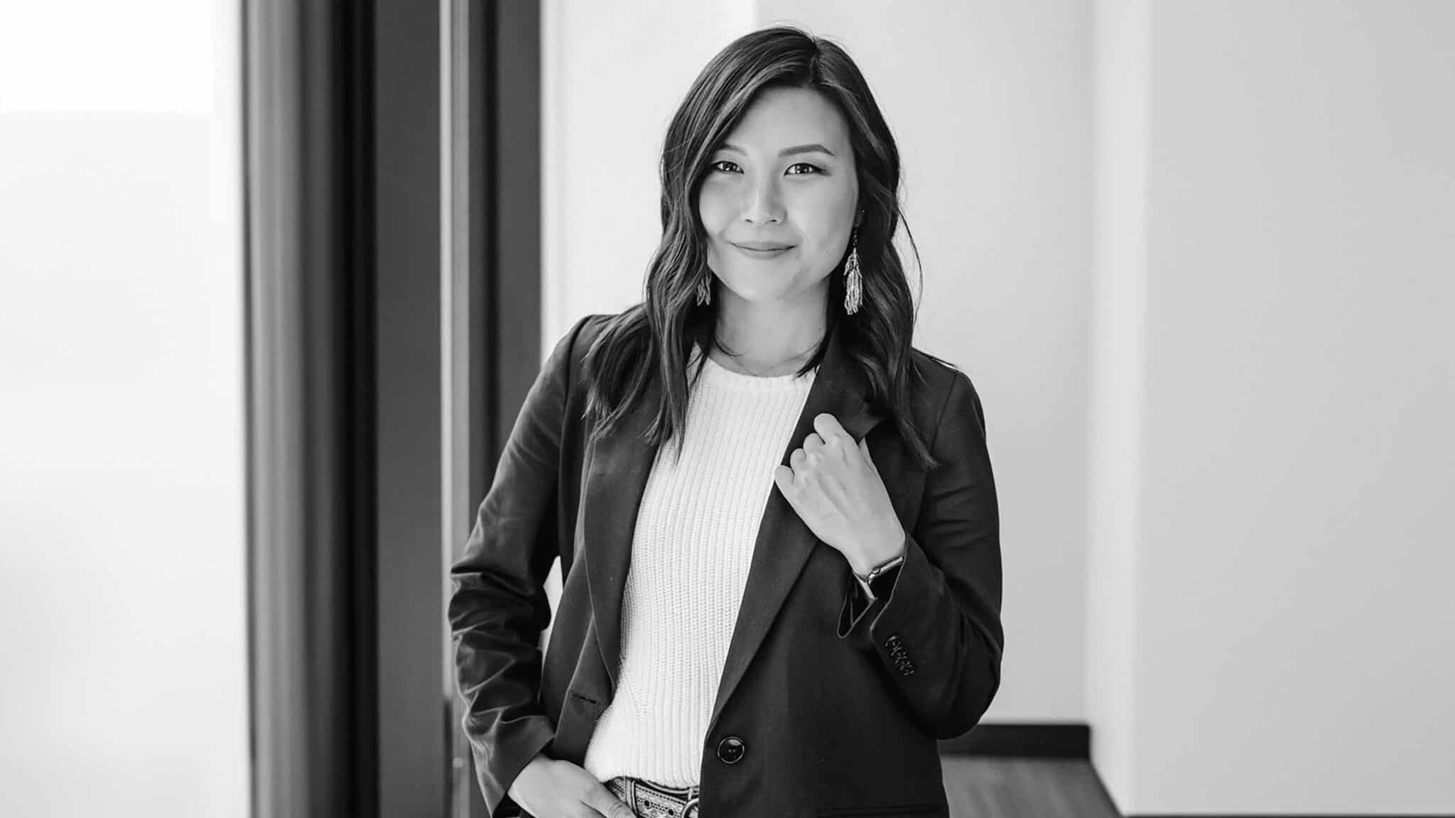 A professional black and white portrait of an asian woman smiling, standing in an office environment, dressed in a blazer and white top.