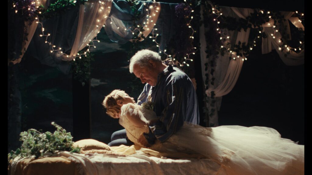 An older man holds a younger woman in a white dress as they sit on a bed surrounded by decorative lights and draped fabric.