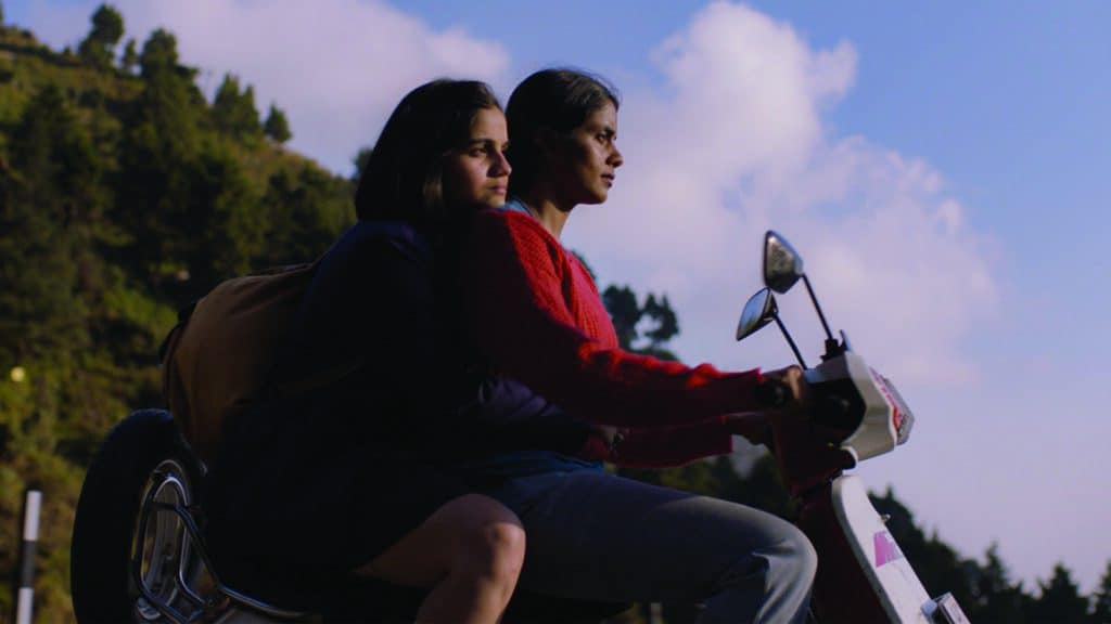 Two people ride a scooter together through a hilly area under a clear sky. The person in front wears a red sweater, and the person in back clutches a backpack.