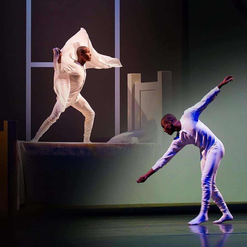 A dancer in white performing on stage with a reflected image on a backdrop creating a symmetrical visual effect, featured in the March 21-27 Must List.