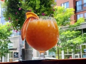 A chilled orange cocktail garnished with a slice of orange, served in a glass on an outdoor table with Seattle magazine in the background.