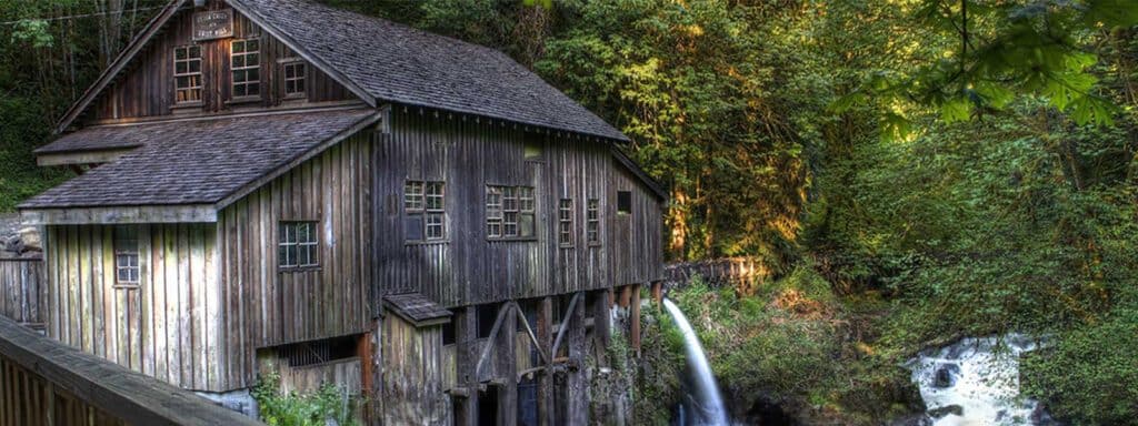 An old wooden mill with a waterwheel situated beside a lush forest in Hidden Washington, with water flowing into a stream.