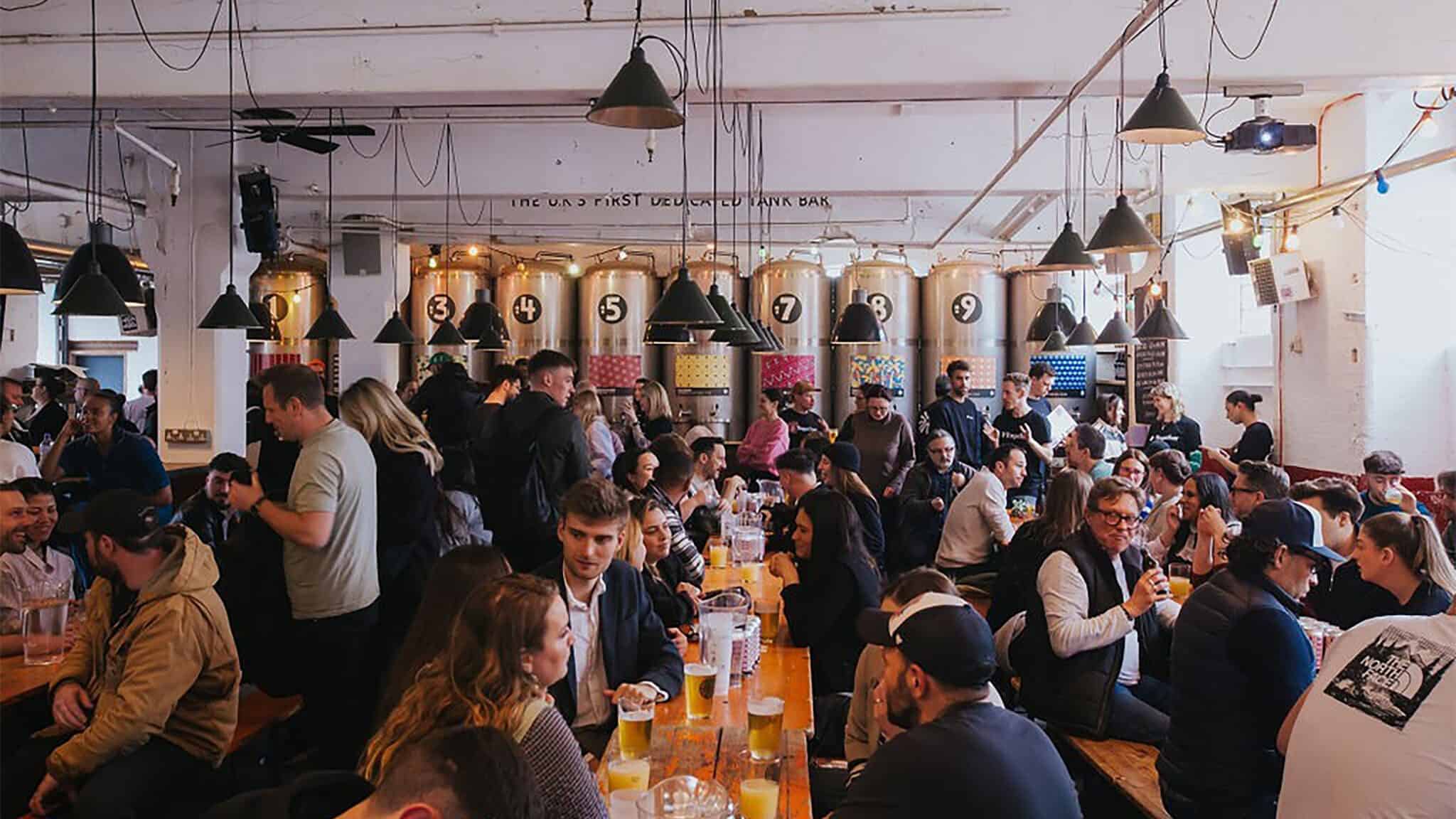 A busy brewery in Pioneer Square with many people sitting at communal tables, some standing at the bar, beneath hanging industrial lights.