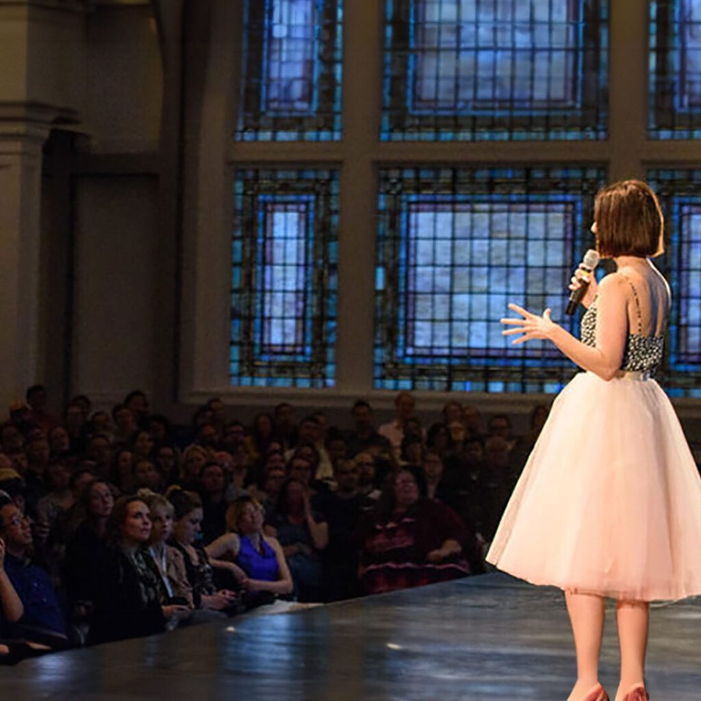 A woman speaking to an audience in an indoor setting with large windows, featured on the Must List for March 21-27.