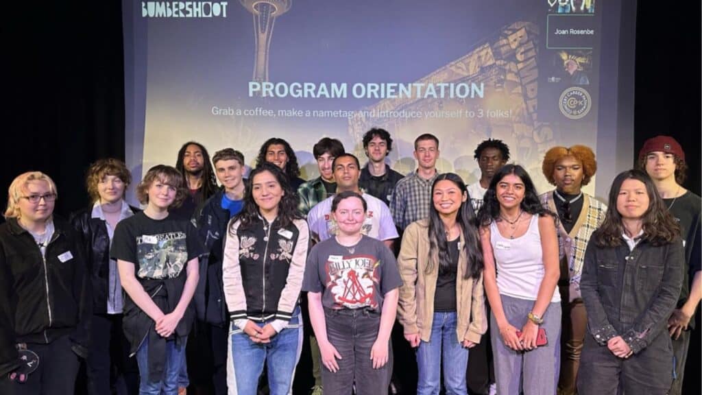 Group of teenagers standing on a stage with a "Bumbershoot program orientation" banner in the background at an event.