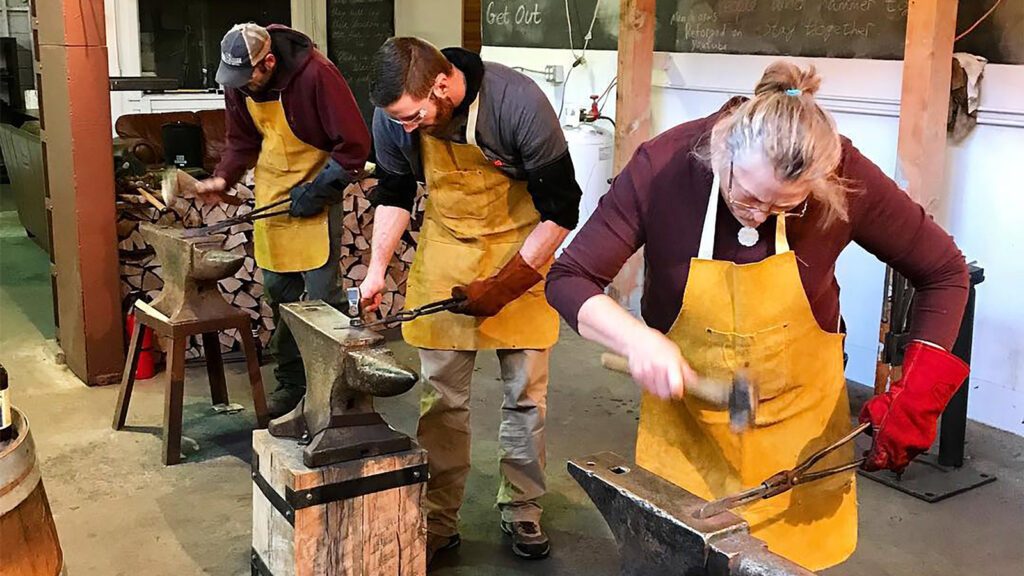 Three friends working with metal on anvils in a blacksmithing workshop during their group getaway.