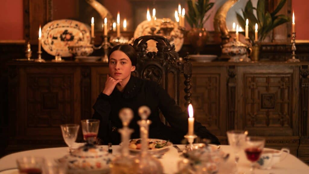 A woman dressed in dark clothing sits at a lavishly set dinner table with various drinks and food, surrounded by ornate decor and lit candles in a dimly lit room.