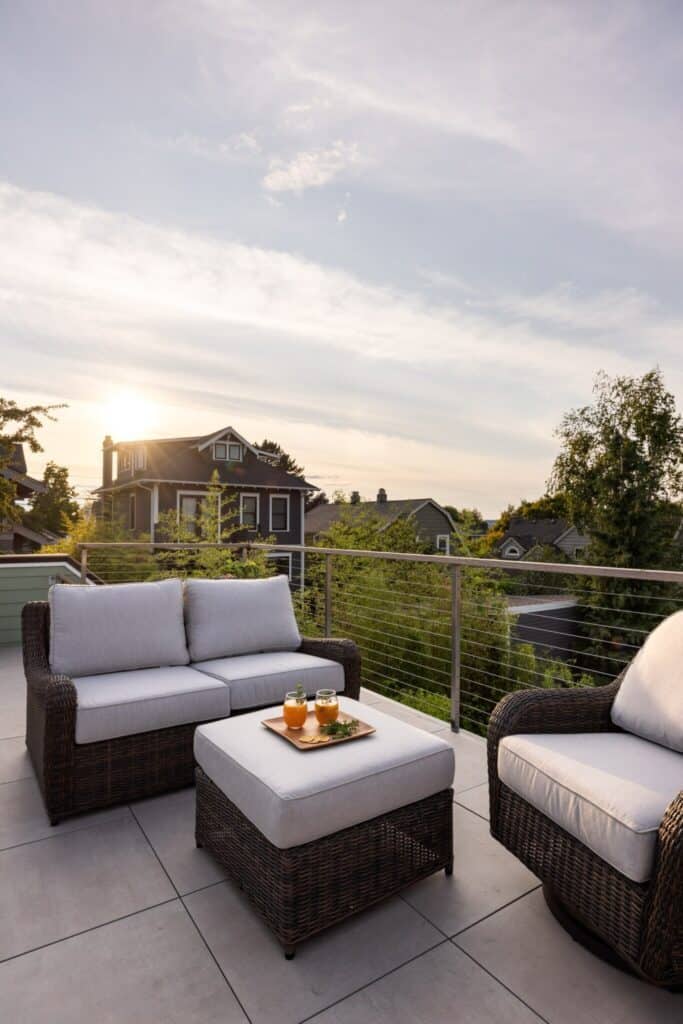 View of the house roof, there are other houses in in the front and 2 garden sofas with a smaller square bench used as a table, where a golden square tray holding two juicy cocktails sits.The furniture is made of dark rattan with light beige cushions. 