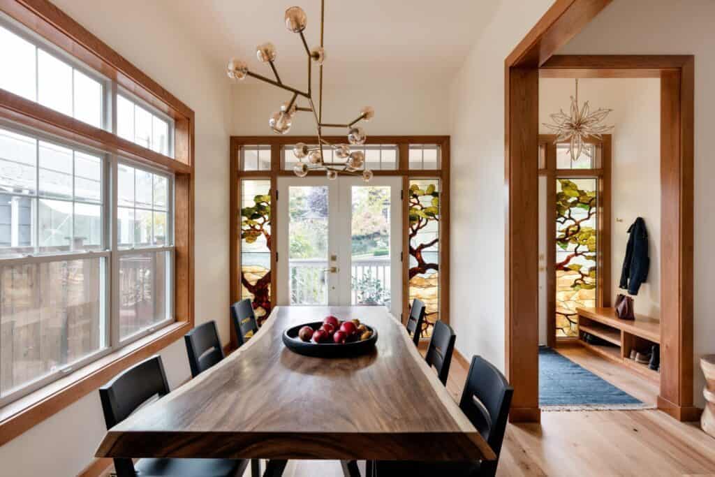 Photo of the dinning room, showing a warm palette. There is a large warm-color wood long table with 3 chairs on each side. The table holds a tray with red apples. At the back is a framed window with 2 doors that look at the front porch.