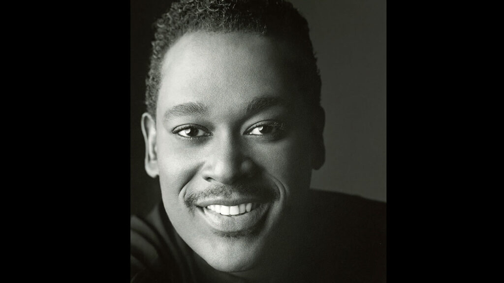 Black and white portrait of a person smiling and looking at the camera. They have short, curly hair and a mustache, reminiscent of classic films.