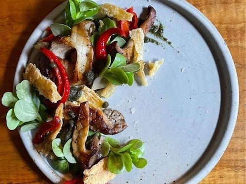 A plate with a mixed salad of leafy greens, sliced mushrooms, roasted red peppers, and crispy bread pieces, garnished with a drizzle of sauce, perfect for enjoying at one of the cozy Sheringham restaurants, on a wooden table.