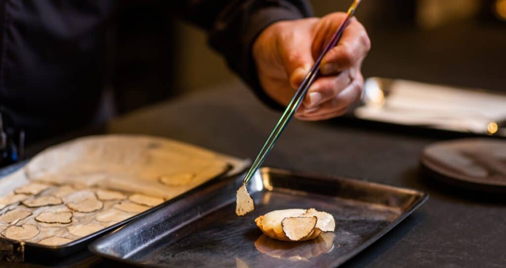 A chef uses tweezers to delicately plate thinly sliced truffles on a farm table, with a tray of more truffles nearby.